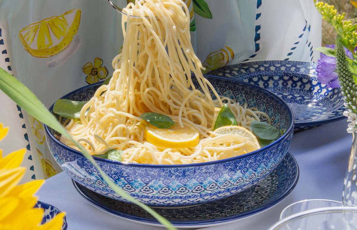 Schaal Porto: Vrouw serveert pasta uit een grote blauwe Bunzlau Castle-schaal aan een zomers gedekte buitentafel.