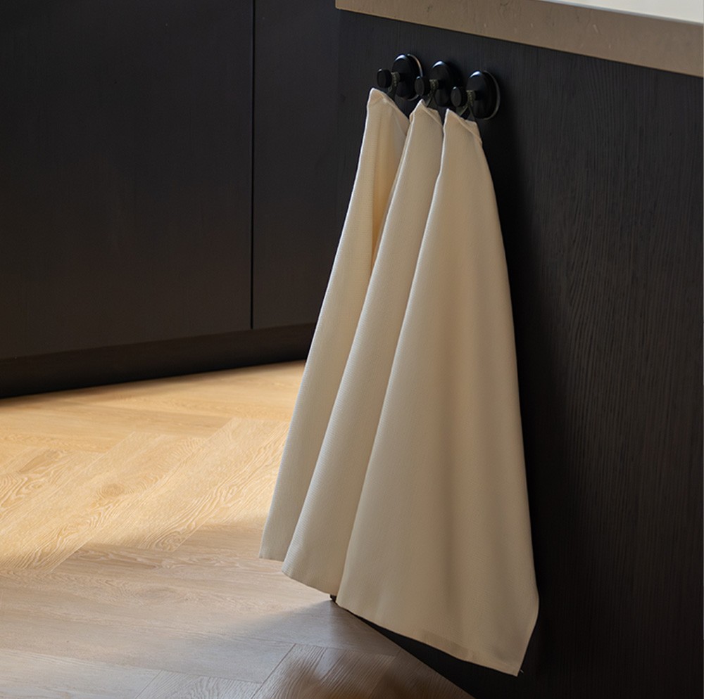 Deux torchons de cuisine couleur crème de la marque Organic Home sont suspendus à un crochet dans une cuisine noire et sombre. Organic Home est un label de Bunzlau Castle.