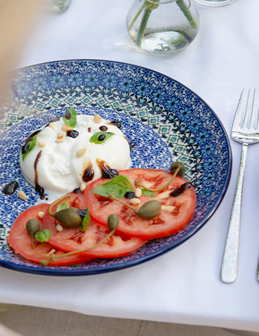 Bunzlau Castle bord in decor Lisboa met tomaat, burrata, kappertjes en basilicum op een zomers gedekte tafel.